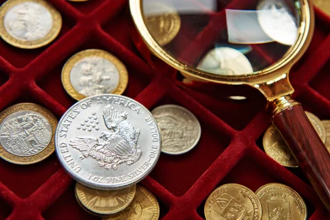 A silver dollar and assorted coins in a collector's tray beside a magnifying glass